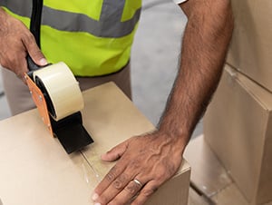 packaging supplies - a warehouse worker taping a shipping box wearing safety vest